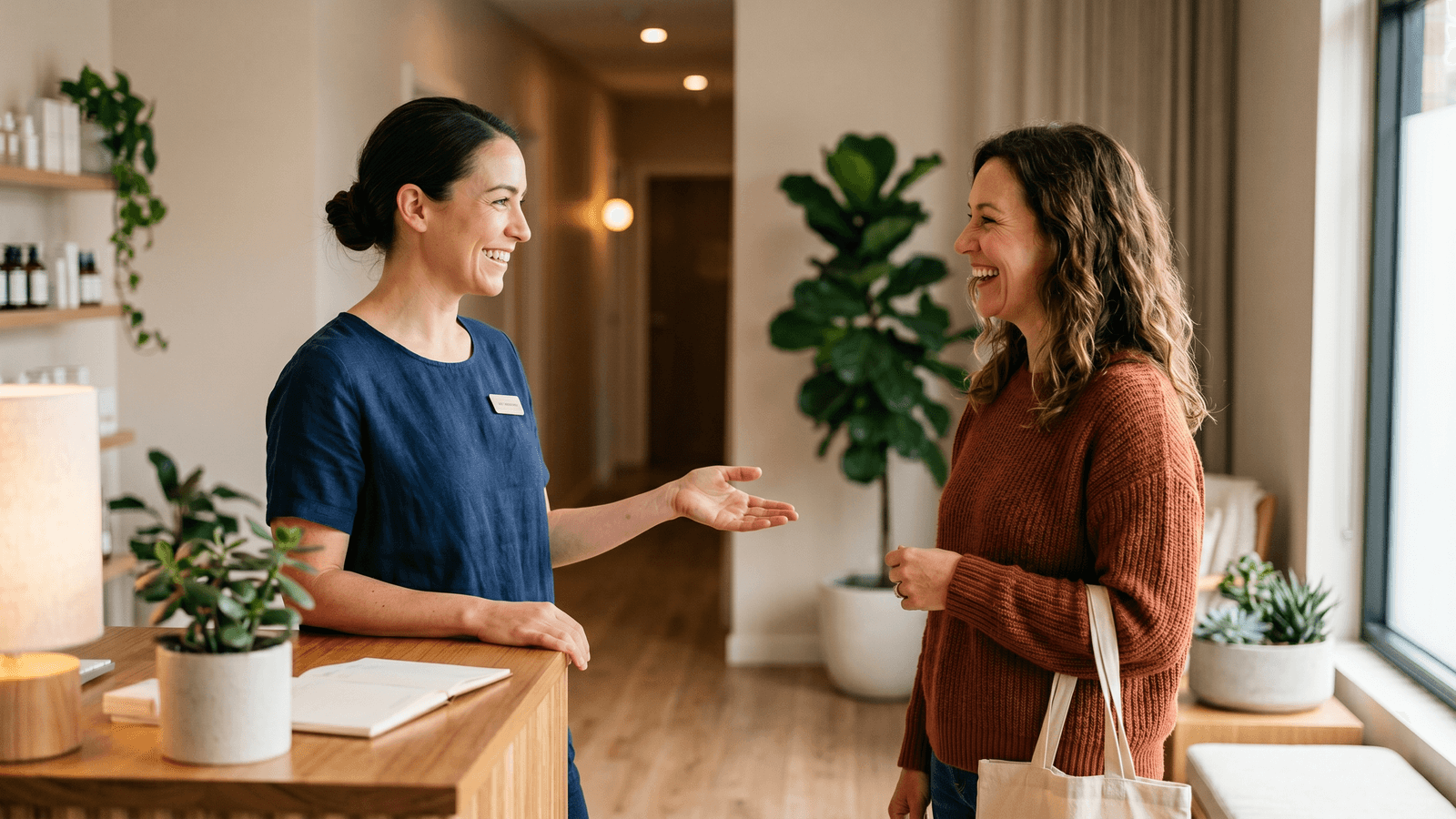 Massage therapist greeting a returning client warmly at a clinic front desk in calm daytime light